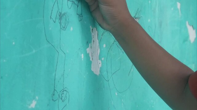 A child's hand holding a pencil and drawing creative scribbles on a green wall surface.