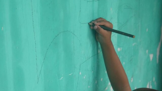 A child's hand holding a pencil and drawing creative scribbles on a green wall surface.