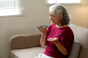 Asian older woman voice command on smartphone speaking at home living room couch modern technology senior communication casual lifestyle activity relaxed retirement
