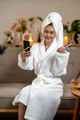 Asian adult woman taking selfie peace sign using smartphone after bath spa treatment at home enjoying cozy relaxation wellness beauty lifestyle