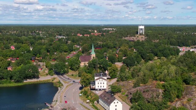 Scenic aerial of traditional church with green spire and modern water tower amid lush forest and residential area in Gustavsberg