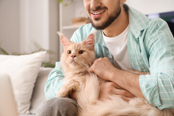 Young man with cute Maine Coon cat sitting on sofa in living room, closeup