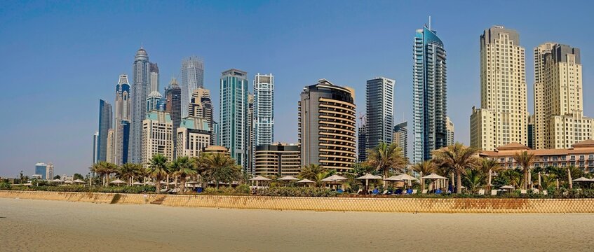 A scenic view of a white sandy beach lined with palm trees, set against a backdrop of towering modern residential skyscrapers under a blue sky.