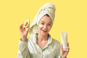 Happy young woman with tube of facial mask and fresh avocado on yellow background