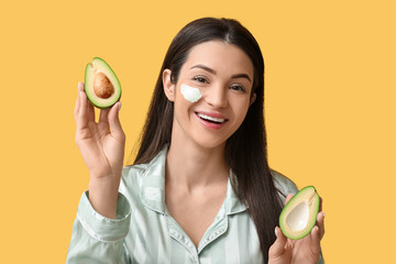 Happy young woman with smear of facial mask and fresh avocado on yellow background