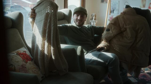 Young man sitting in armchair, looking sad while holding up two coats. Moment reflects emotional decision or winter clothing choice.
