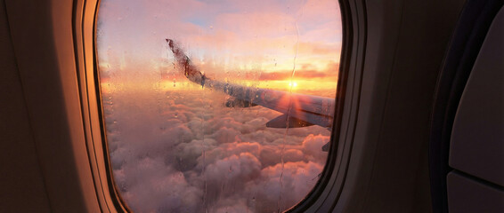 Airplane wing over clouds at sunrise reflecting sun flares on window pane
