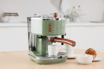 Modern green coffee machine with bowl of sugar, cups and cupcake on table in kitchen