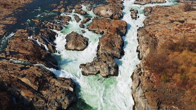 Drone shot of the powerful Khone Falls cascading over rugged rocks in southern Laos. The turquoise water flows through dramatic rocky terrain, creating a stunning natural landscape.