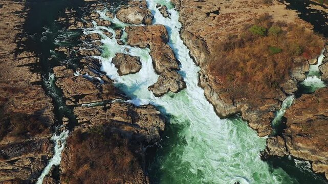 Dramatic aerial shot of Khone Falls with powerful rapids flowing between rocky cliffs and sparse vegetation in Laos.