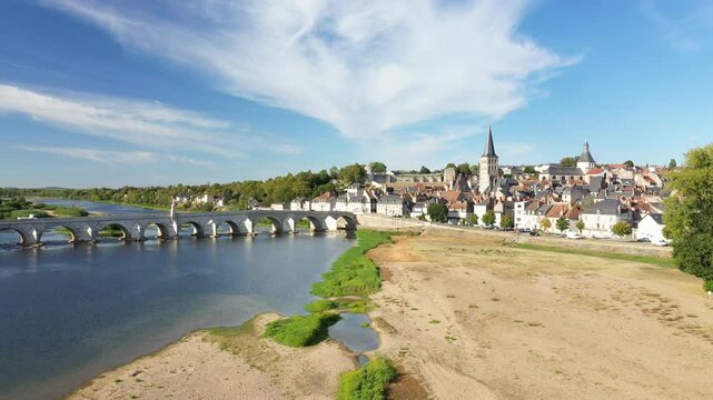 Aerial view of La Charite sur Loire with its historic stone bridge spanning the Loire River under a blue sky. Picturesque old town architecture and riverside landscape are visible.