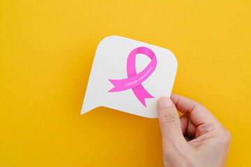 hand holding real white speech bubble paper cut with pink ribbon icon and cancer awareness text on yellow background