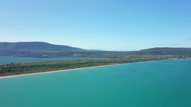 Drone shot of the turquoise sea and lush coastline at Isola Varano Nature Reserve in Italy, with mountains in the background under a clear blue sky.