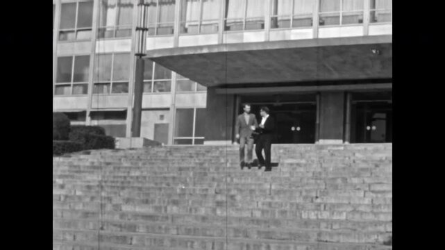 Two men in suits walk down stairs on office building backdrop. Smiling businessmen, confident entrepreneur chat about job in morning city center. Success in old film. Archival retro. Vintage archive