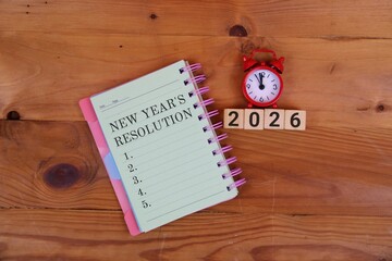 a spiral notebook labeled New Year’s Resolution, wooden number blocks spelling 2026, and a small red alarm clock, suggesting planning, goals, and a fresh start for the new year.