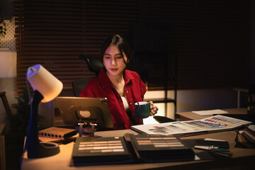 Interior creative designer working overtime at night in a cozy home office, reviewing a draft on a tablet while holding a cup of coffee