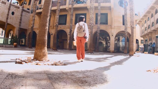 Rear view. A carefree female tourist in a straw hat, white shirt, and backpack walks along an old street. Jaffa. Israel. Tourism and travel concept.
