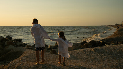 Two children, a boy and a girl, holding hands and walking together on a rocky beach towards the ocean during a warm sunset, symbolizing family connection, childhood, and summer freedom.