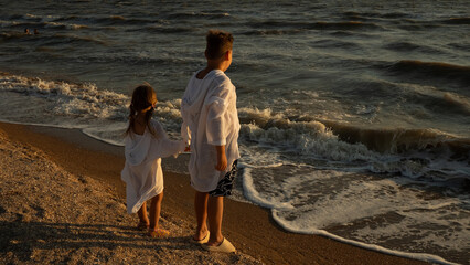 Siblings standing on a sandy beach holding hands, observing the gentle waves of the sea washing ashore during a warm evening sunset, capturing a moment of childhood togetherness and connection.