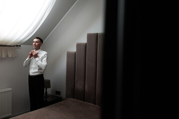 Groom prepares for wedding in cozy room, adjusting his shirt and tie before the ceremony on a sunny day