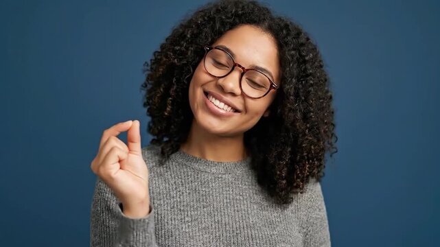 A cheerful young woman with a radiant smile and stylish eyeglasses gestures with her fingers, indicating a small amount or size against a clean, minimalist blue background. Her expressive facial featu