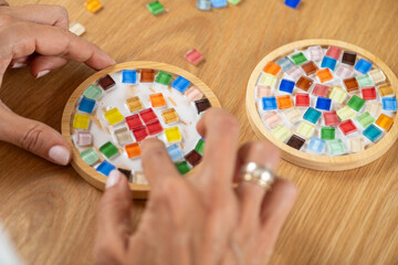 Making mosaic coasters at home as a hobby. Arranging colorful glass parts and gluing them onto coaster bases. Shows material awareness, careful alignment, and deliberate creative process.