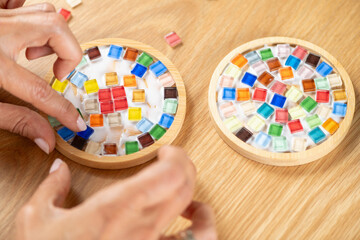 Assembling mosaic coasters at home as a DIY craft project, attaching small moisaic components. Captures focused posture, slow craft rhythm, and mindful artistic attention.