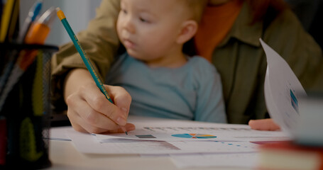 This young mother multitasks by working on her laptop and analyzing graphs while holding her baby. She takes notes and stays organized in her home office.