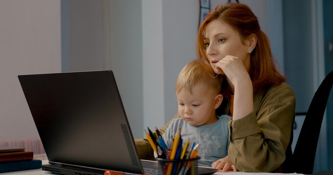 Young mother is multitasking by typing on her laptop while holding her baby. She is focused on her work in a cozy home office environment. - Powered by Adobe