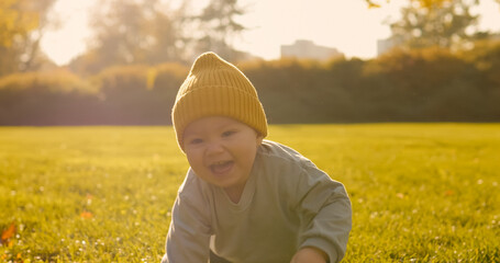Cute infant wearing yellow beanie crawling across lush green lawn, basking in golden sunlight during peaceful park afternoon