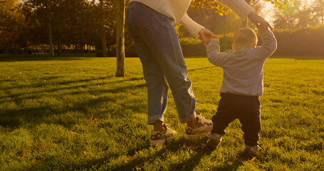 A joyful moment unfolds in the park as a mother encourages her child to take his first steps on the green lawn, basking in the warm sunlight.