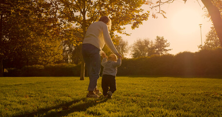 A young boy experiences the joy of taking his first steps in a park, guided by his mother. They enjoy a sunny day on the grass, capturing a moment of growth.