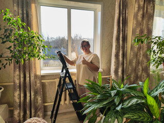 Woman smiling on a stepladder giving a thumbs up after finishing cleaning a sunlit window in a cozy apartment with houseplants, feeling satisfied and accomplished.
