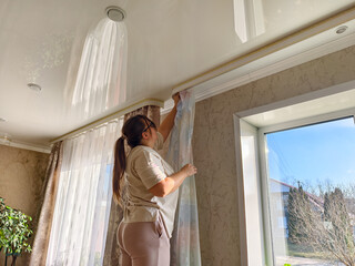 A woman on a stool hangs translucent white curtains on a window ledge. A woman adjusts soft drapes in a bright living room during a DIY home decorating and renovation project.