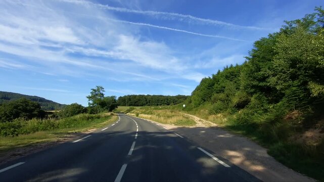 Empty rural road winding through green countryside and forests under a blue sky in the Morvan region of France. Peaceful summer landscape with natural beauty and open space.