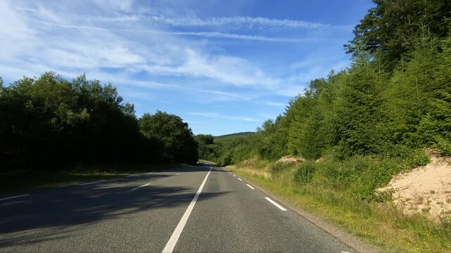 Empty country road winding through lush green forest in the Morvan region of France under a blue sky with wispy clouds. Peaceful rural landscape ideal for travel and nature concepts.