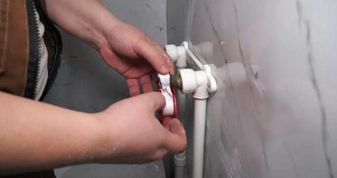 Professional plumber's hands carefully wrapping white sealing tape around the threads of a water pipe fitting to prevent leaks during plumbing repairs or installation in a bathroom.
