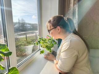 Young woman stands by a bright window holding a blooming geranium, soaking up morning sunlight and calmly gazing out, enjoying a peaceful moment at home.