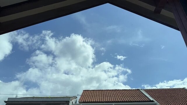 Time lapse A clear blue sky filled with beautiful white cumulus clouds seen from below a sheltered overhang with rooftops in the foreground