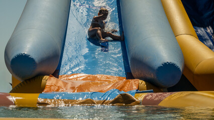 A child slides down an inflatable slide at a water park. The child splashes in the cool water of the inflatable slide and pool, enjoying active summer fun and relaxing in the bright sun.