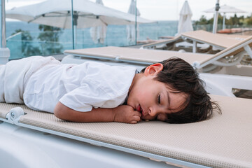 A young boy is sleeping on a lounge chair. The chair is white and has a green tag on it. The boy is wearing a white shirt and shorts