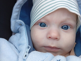 A baby with bright blue eyes looks directly at the viewer while wearing a light blue jacket and a striped hat outdoors on a sunny day. The baby appears attentive and curious