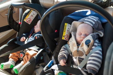 Two infants sit in car seats while secured with safety straps. They appear to be asleep during the car trip. Sunlight enters the vehicle