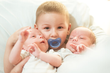 An older brother holds two baby twins in a warm setting. The brothers share a moment of joy, with smiles and a sense of togetherness in their home during the afternoon
