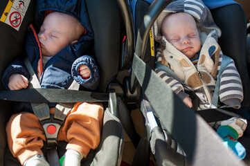 Two babies rest in their car seats while out for a stroll in the park. The bright sunlight shines down, and the children are bundled up in warm clothing
