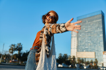 Woman joyful urban lifestyle candid authenticity golden smile as she reaches out across sunlit city plaza in beanie and cardigan, mindful living atmosphere and emotional storytelling vibe.