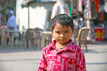 Portrait of a young Indian child with striking light eyes and a serious expression, wearing a red patterned shirt and beaded necklace, standing in a street market area.