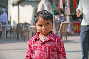 Portrait of a young Indian child with striking light eyes and a serious expression, wearing a red patterned shirt and beaded necklace, standing in a street market area.