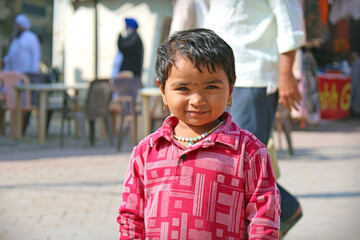Portrait of a young Indian child smiling with striking light eyes and a serious expression, wearing a red patterned shirt and beaded necklace, standing in a street market area.