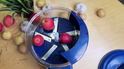 Overhead kitchen shot as a hand loads red apples into a blue fruit and vegetable peeler on a wooden countertop with potatoes and garlic in the background for cooking and meal prep